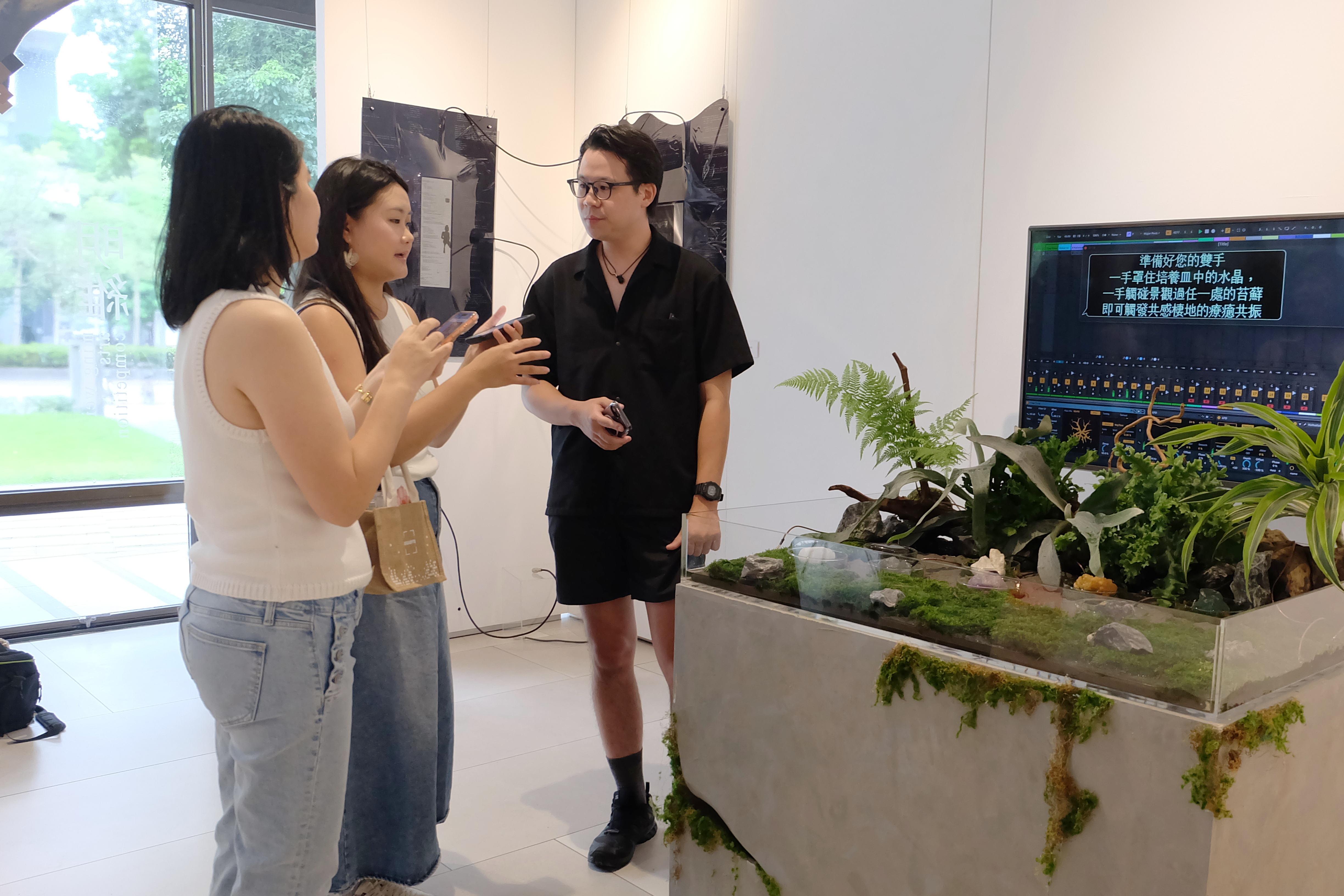 兩位女子與一位黑衣男子在藝廊植物展台旁交流討論。   