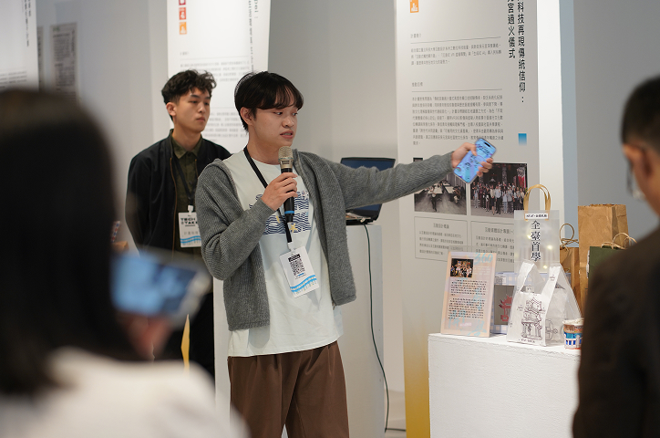 A young man in a gray cardigan holds a microphone and displays his phone screen alongside exhibition items.