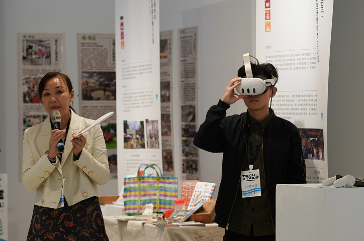 A woman in an off-white jacket speaks into a microphone while a young man beside her wears a white VR headset.   