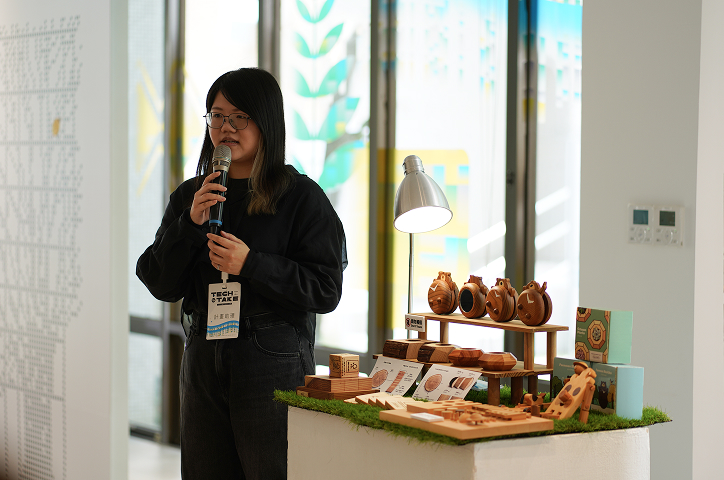 A woman with glasses wearing a black top holds a microphone and introduces wooden craft items on display.
