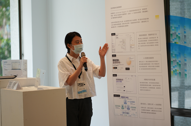 A woman wearing a mask and a white short-sleeved shirt holds a microphone and gestures toward an information board.
